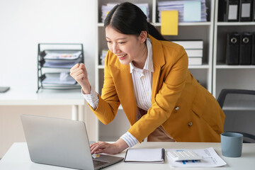 Young Asian businesswoman in a yellow suit showing joy through a laptop and smartphone to colleagues for accomplishing a company project with set objectives, the concept of success.