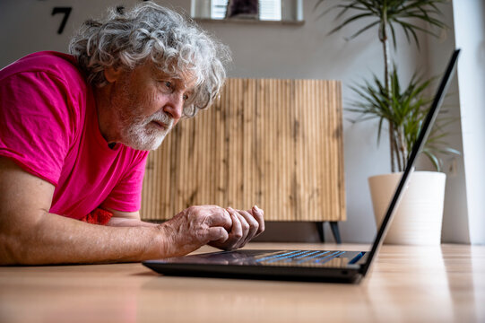 Senior man in pink shirt lying on the living room floor typing on laptop computer