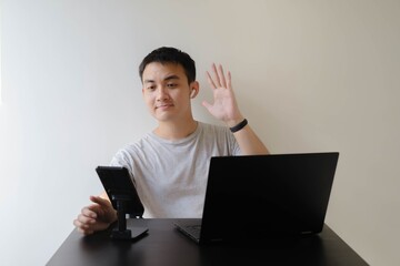 A young Asian man wearing a grey t-shirt, a smartwatch on his left wrist, and a pair of wireless earphones is waving his hand at people at an online meeting. Isolated white background.