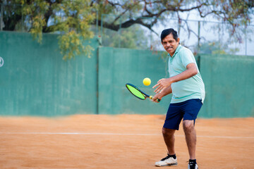 Excited indian senior man busy playing tennis by hitting ball with racquet and court - concept of...