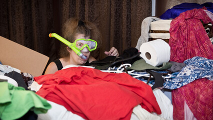Swimming in Stuff. Wearing a snorkel and goggles an overwhelmed woman with messy hair dives into a large pile of miscellaneous clothes and household items. Copy space.
