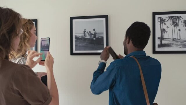 Group of visitors taking pictures of framed photo on wall while visiting exhibition in art gallery