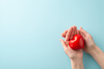 First person top view photo of female hands cradling a red heart on a pastel blue background....