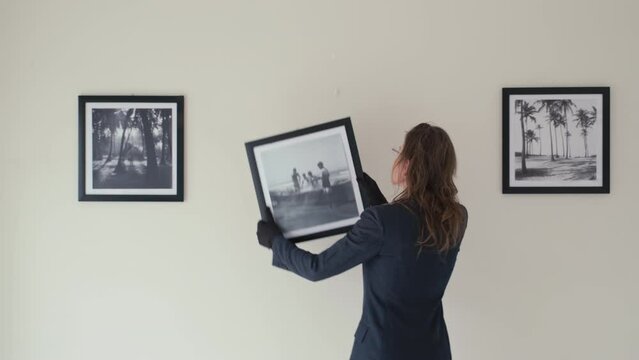 Female gallery curator in gloves hanging framed photo on wall, looking at it and fixing while preparing exhibition hall