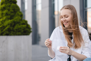 Happy blonde woman sitting on the bench near office and take out white wireless earphones from headphones case. Girl want listen music or talk by mobile phone using earbud. Happy business girl.