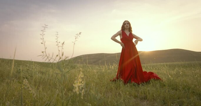 Slow motion, professional female model posing at golden sunset. Professional fashion model in long red dress at sunset in field. Concept of haute couture, high fashion, exquisite style and vogue.