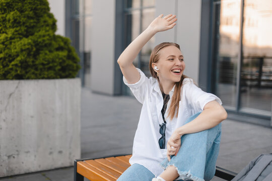 Happy Blonde Woman Make Hi Gesture, Wave Her Hand While Meet Colleagues Near Office Building, Sitting On Bench And Listen Music In Wireless Earbuds. Goodbye Or Hello Gesture Of Business Woman.