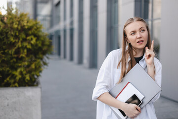 Blonde businesswoman with laptop and book in hand, have idea. point finger up, wow emotions outside office background. Concept of idea and brainstorm. Woman have new ideas or search solutions.