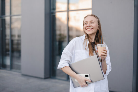 Beautiful woman going to work with ice latte walking near office building. Successful business blonde girl holding cup of hot drink, laptop, phone on her way to work on city street. High resolution.