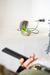 hearing aid for deafness on charger on top of white dining table, detail of unrecognisable eighty year old man's arm and hand on the sofa with the TV remote control.