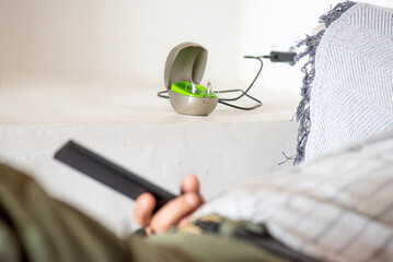 hearing aid for deafness on charger on top of white dining table, detail of unrecognisable eighty year old man's arm and hand on the sofa with the TV remote control.