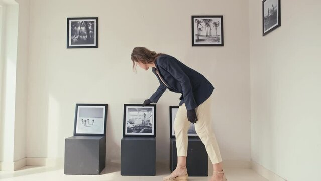 Full shot of female gallery manager in gloves choosing framed photo and putting it on wall while preparing art exhibition