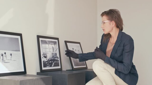 Experienced female gallery curator in gloves choosing artwork and putting frame on wall while preparing photo exhibition