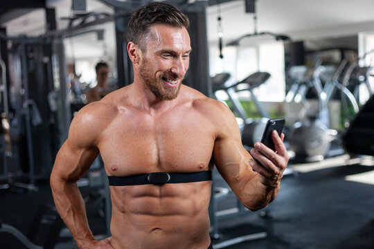 Strong, fit, athletic man in a gym with heart rate monitor around chest checking heart rate on cell phone with a big smile. Good result, good training