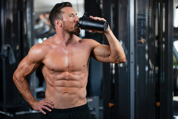 Fototapeta na wymiar Strong, fit, athletic, attractive man in a gym. Drinking protein or water from the black shaker .