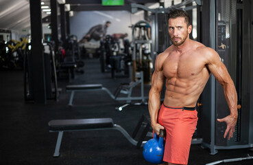 Fototapeta na wymiar Strong muscular man standing in a gym with blue kettlebell in one hand.