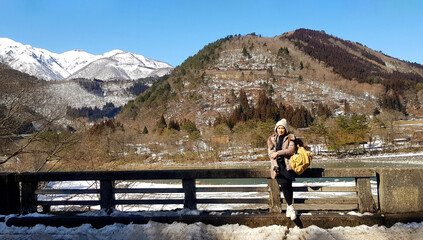 Asian tourist woman in brown coat and yellow knit hat standing on bridge with tree, snow on mountain and blue sky background with copy space at Japan. People travel in Asia with natural and landmarks.