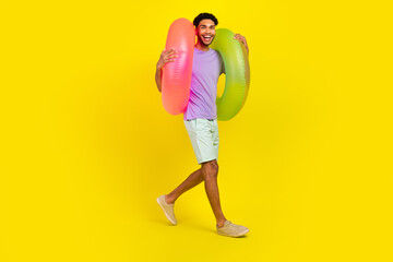 Full size body cadre of young guy enjoy pool parties walking with inflatable rings lifebuoy beach tour isolated on yellow color background