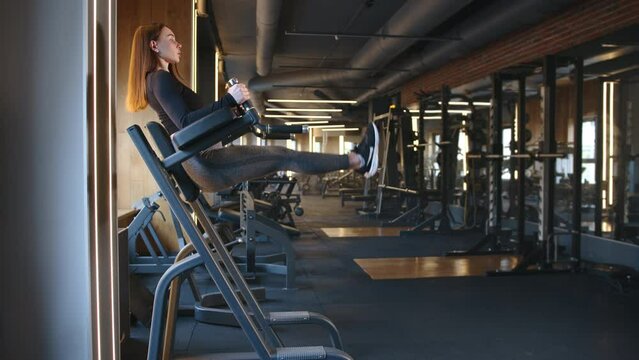 Young fit woman performing vertical leg raise for press in fully-equipped gym. Woman raises straight legs, forearms on the padded bars, body is straight. High quality 4k footage
