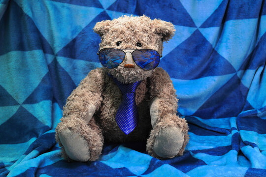 Cute Teddy Bear wearing a tie and sunglasses sits on a blanket with a blue triangle geometrical pattern. Charming businessman concept.