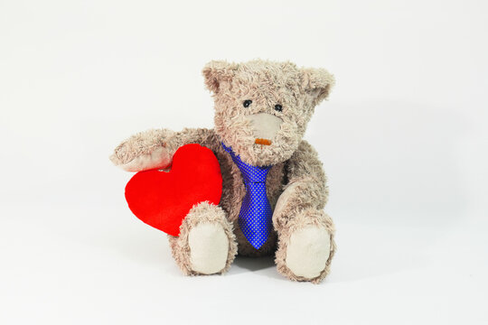 Charming plush Teddy Bear wearing blue necktie sits on white photographic background and holds a red heart under his hand. Charming, cute and elegant concept.