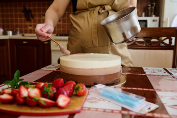 Close-up of female confectioner decorating a triple chocolate mousse cake with ruby pink icing in the home kitchen Sliced fresh strawberries on the cutting board. Food still life. Making birthday cake