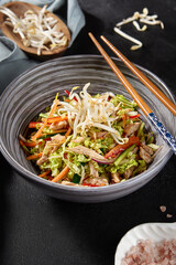 Side view vertical shot of a delightful chicken dish in an Asian style bowl. It's made with vegetables, Peking cabbage, soy sprouts, and a touch of pink sea salt, served on a black