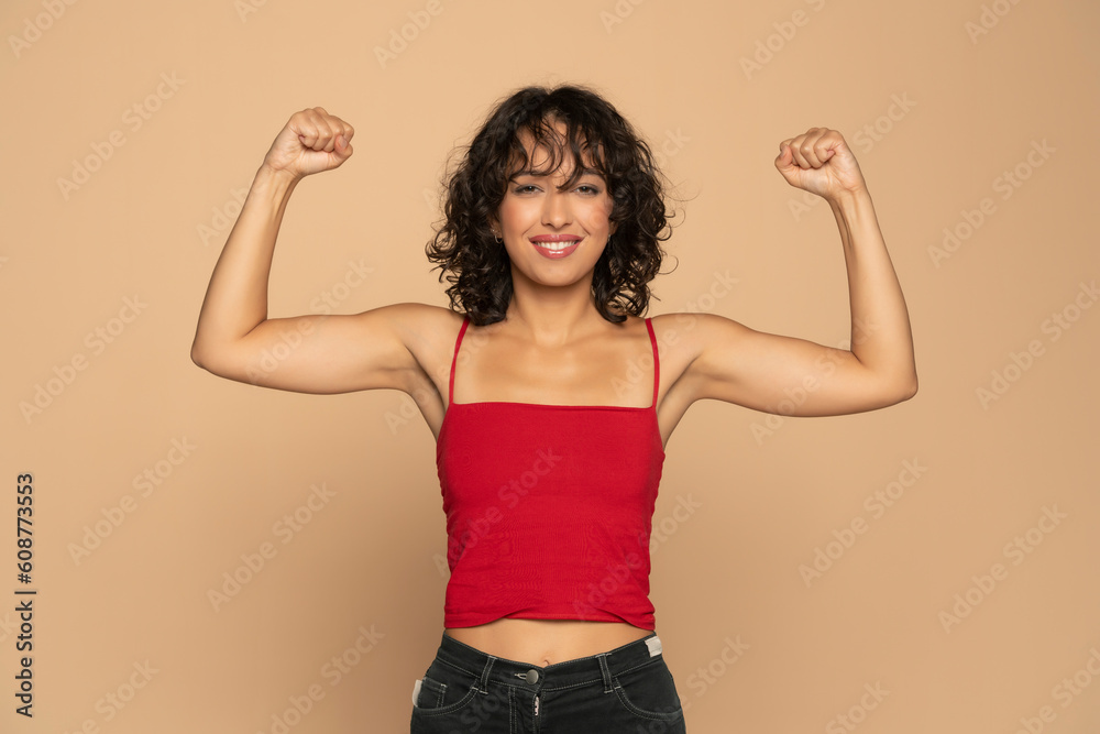 Wall mural young exotic happy brunette woman in red sleeveless shirt and jeans showing biceps on a beige backgr - Wall murals