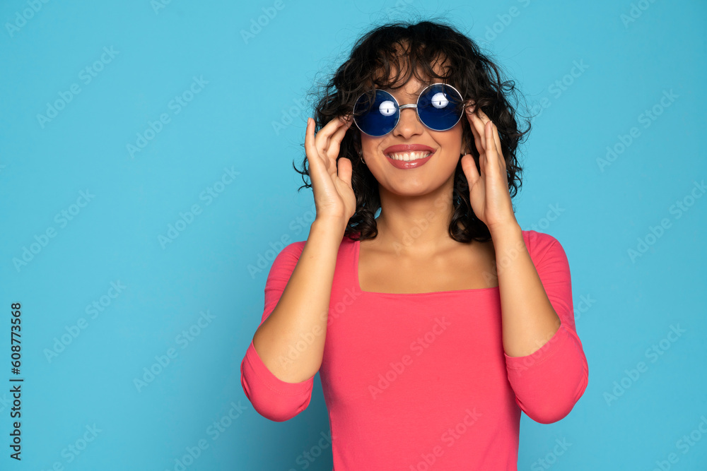 Wall mural Young exotic smiling brunette woman in pink blouse posing with a sunglasses on a blue background