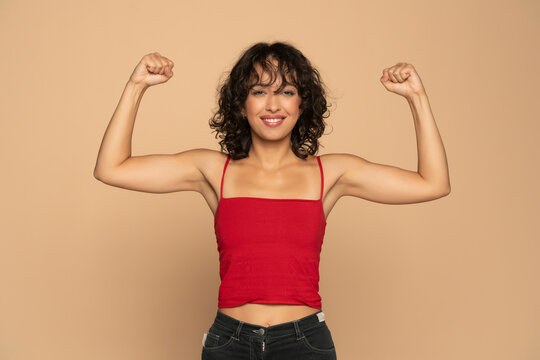 Young exotic happy brunette woman in red sleeveless shirt and jeans showing biceps  on a beige background