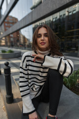 Fototapeta na wymiar Fashionable beautiful young woman model in a stylish striped sweater sits and looks at the camera in the city