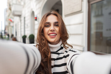 Happy beautiful young girl with a cute smile in a fashionable striped sweater makes a selfie photo on a smartphone and travels in a European city