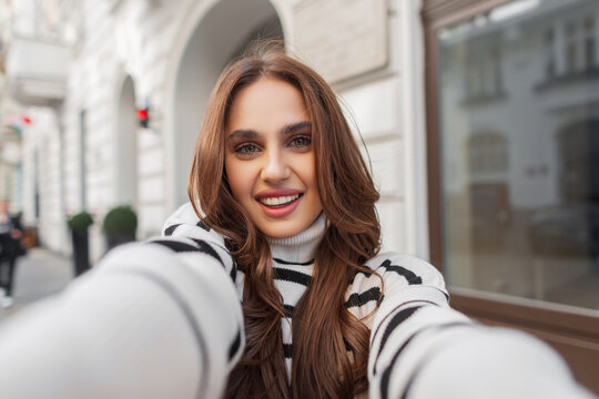 Happiness young pretty woman with cute smile in fashion striped sweater enjoy travel and make selfie on smartphone near a white vintage building in the European city