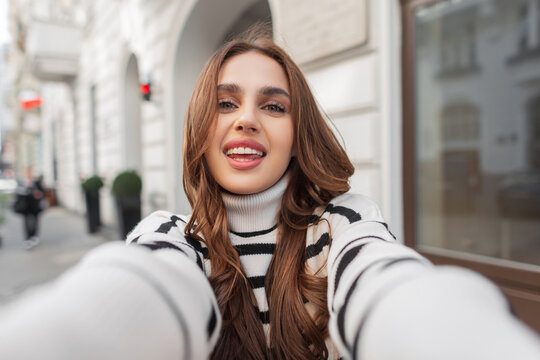 Funny beautiful young woman with a white cute smile in a trendy fashion striped sweater travels and takes a selfie photo in the city near a vintage building