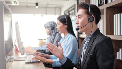 Young Asian male customer support phone operator with headset working in call center. Young man using headset and computer in modern office