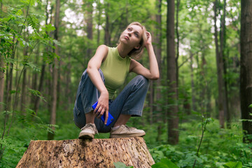 teenage girl sits on a stump in a natural forest park, thinking and holding a smartphone in her hand