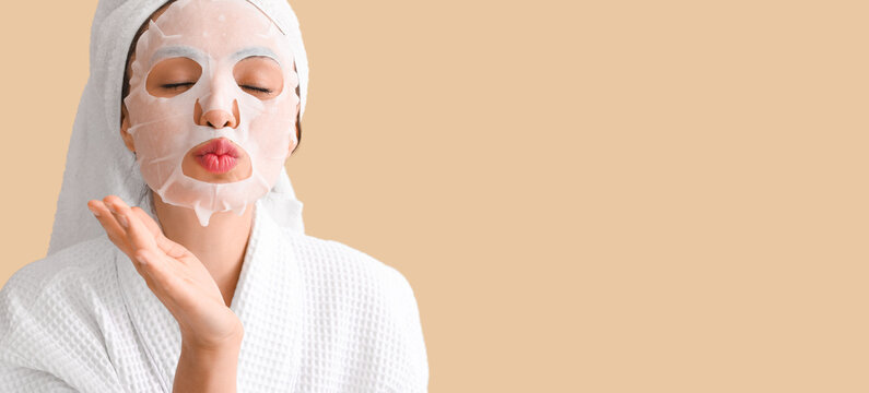 Young woman with applied sheet facial mask blowing kiss on beige background with space for text