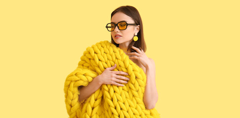 Stylish young woman with knitted plaid and sunglasses on light yellow background