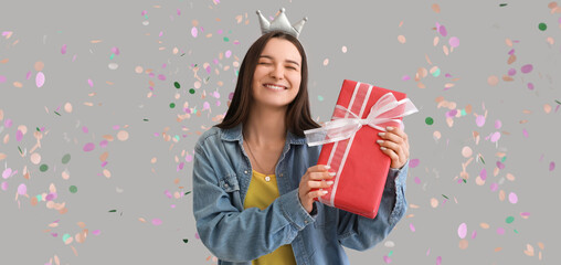 Happy young woman with birthday gift and falling confetti on grey background