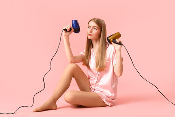 Thoughtful young blonde woman in pajamas with hair dryers on pink background - obrazy, fototapety, plakaty