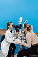 Child doctor ophthalmologist with slit lamp in medical coat. Child is looking through slit lamp and...
