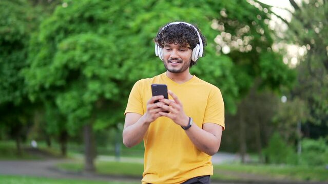 Young smiling active guy sporty walking with headphones mobile phone enjoying listening to music, jogging outdoors on urban city park or forest background. Cheerful runner in t-shirt on training