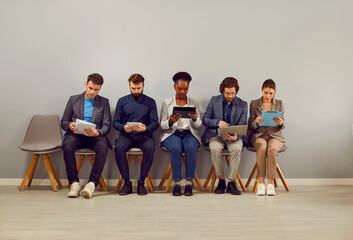 Group of diverse multiracial people sitting in line on chairs by gray office wall, waiting for work...