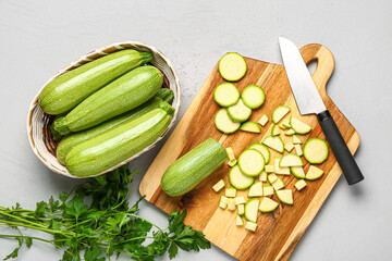 Wicker bowl with many fresh green zucchini and board of slices on grey background