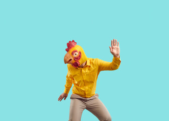 Happy guy in yellow chicken mask having fun at crazy party. Funny cheerful carefree young man wearing bizarre bird mask dancing isolated on turquoise background