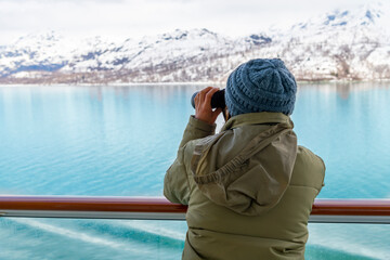 A young woman with binoculars views the snow covered mountains and glaciers from a cruise ship balcony at Glacier Bay National Park and Reserve, Alaska USA. - obrazy, fototapety, plakaty