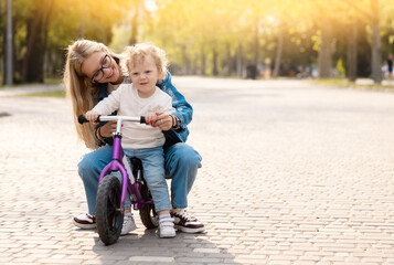 Young mother teaches her little son how to ride a balance bike in the park