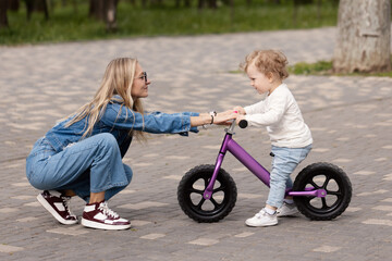 Young mother teaches her little son how to ride a balance bike in the park