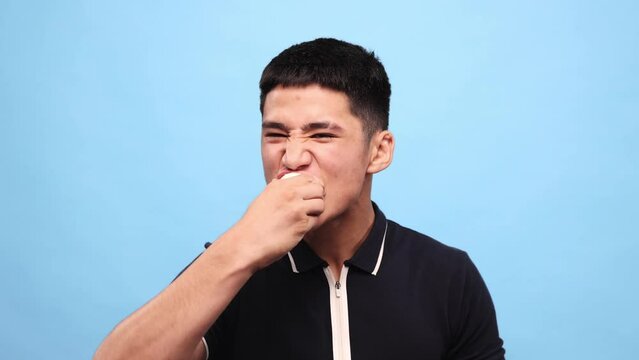 Young asian guy biting onio against blue studio background. Handsome young man with grimacing face eating spicy, bitter vegetable. Concept of taste, human emotions, facial expression