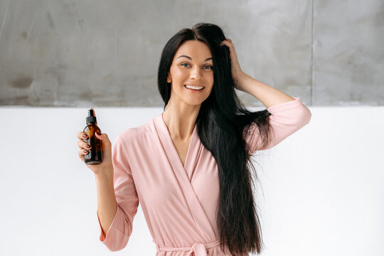 Lovely brunette caucasian woman applying essential oil spray on her long hair while sitting in a bathroom, looks at camera, smile. Split ends repair treatment. Female beauty routine. Haircare concept